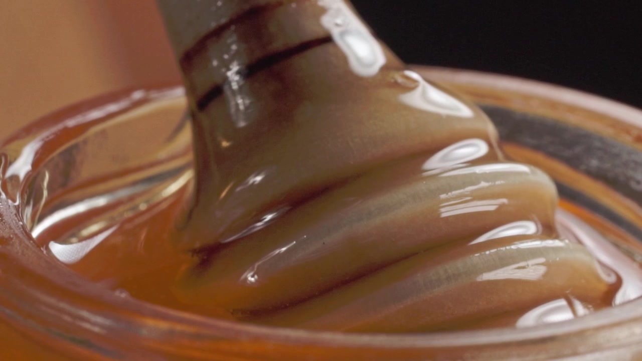 Macro close up of thick amber colored syrup with a glossy texture being stirred in a glass container, showing smooth folds and viscous consistency