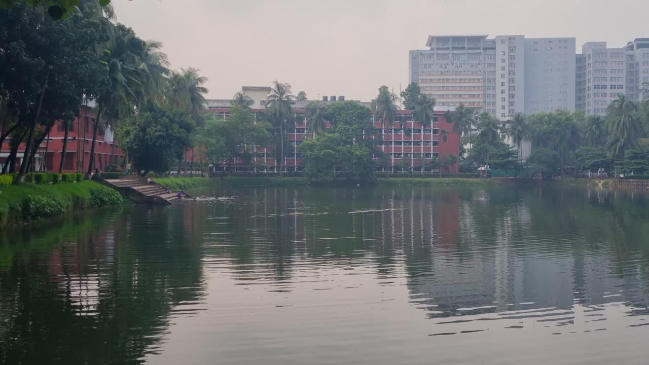 Vista dello stagno di Shahidullah Hall presso l'Università di Dacca, in Bangladesh, incorniciato da palme ondeggianti ed edifici storici in pietra rossa, sotto un cielo velato e nebbioso