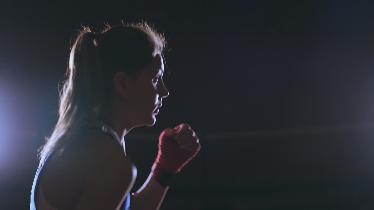 A beautiful sports boxer woman in red bandages on her hands and a blue t-shirt is fighting with a shadow practicing the speed and technique of punches. Camera movement side View. Steadicam shot. Preparing for self-defense