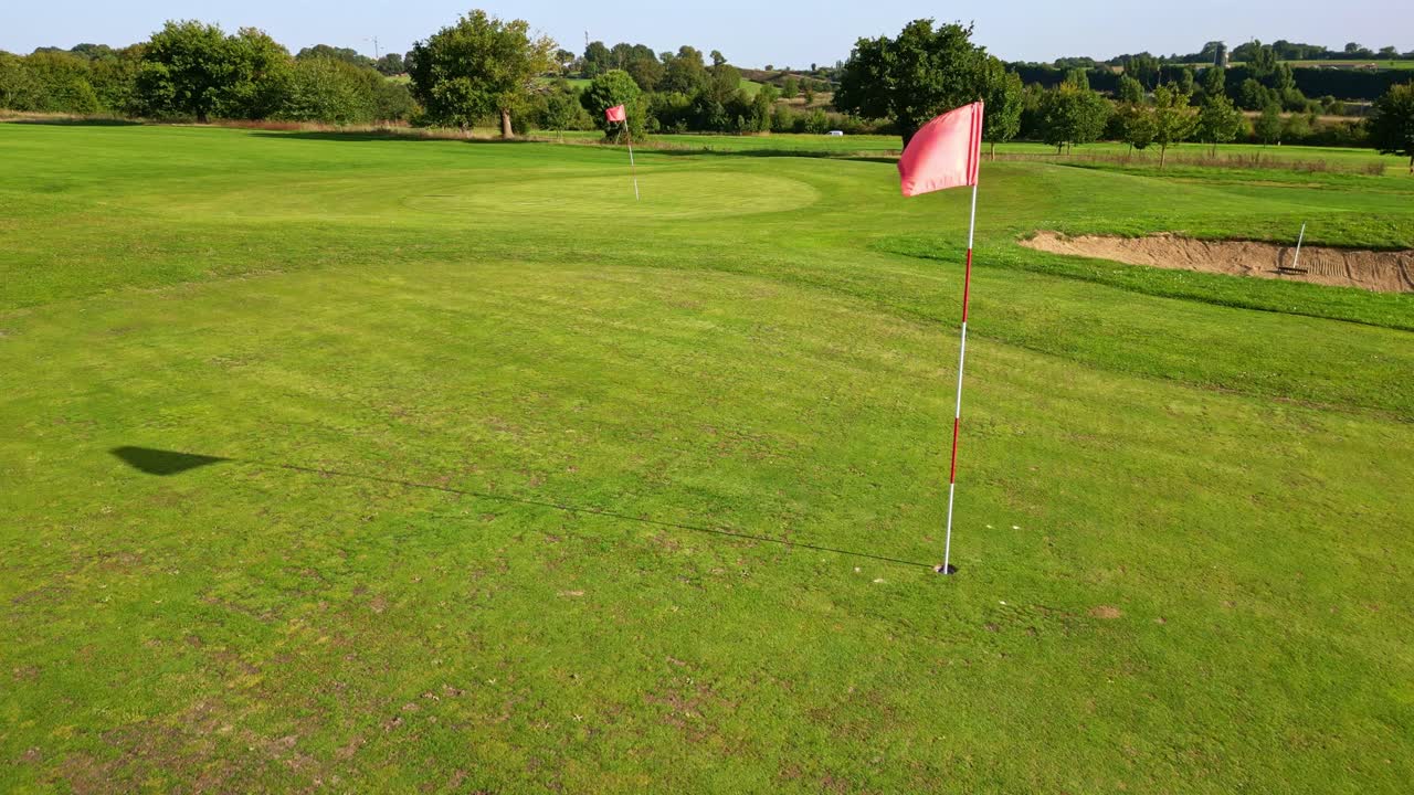 Drone flies low across a golf course, passing close to a flag and heading toward another in the distance, with grass areas, trees and a car visible