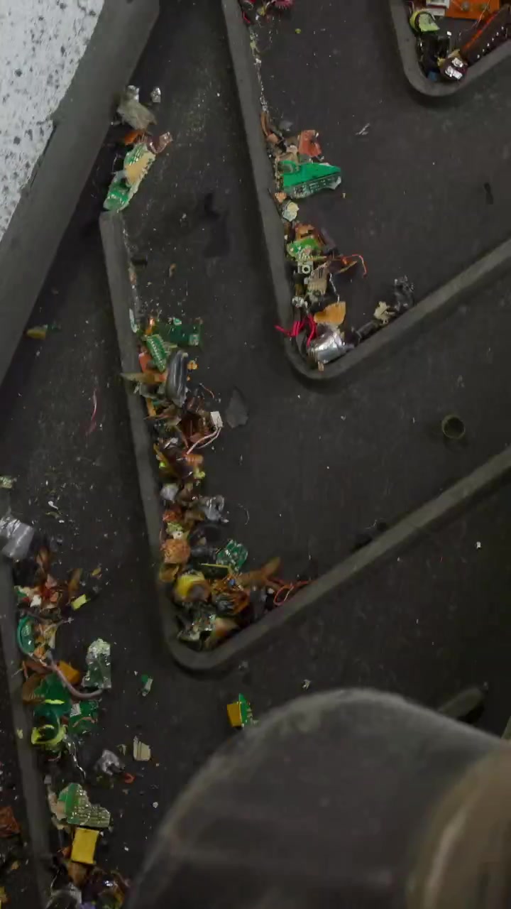 Primo piano di nastro trasportatore che trasporta frammenti di plastica, con piastre metalliche a spina di pesce e scaglie multicolori sparse.