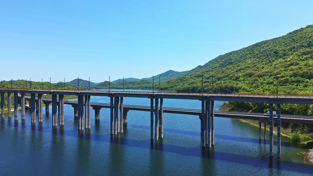 Vista de un puente largo sobre el agua rodeado de colinas verdes bajo un cielo azul durante el día, mostrando la carretera y las vías del tren.
