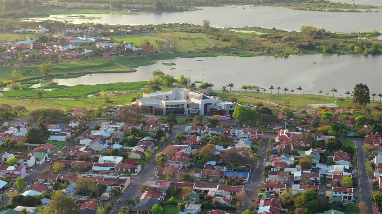 Aerial shot of a river beside an urban area with a prominent building amidst red-roofed houses, showcasing a harmonious blend of natural and man-made elements in Federacion, Entre Ríos, Argentina