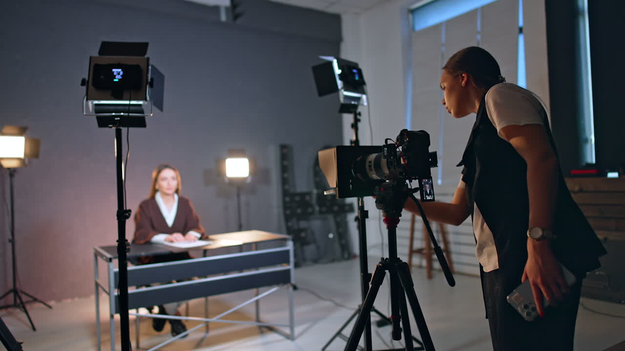Female photographer fixes camera directed on the woman sitting at desk. Blog footage in studio.