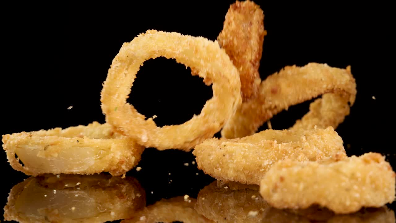 Golden fried onion rings fall and bounce onto a glossy black surface under bright studio lighting, captured in slow motion with a static camera angle