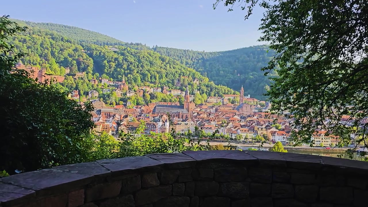 Vista panoramica su Heidelberg, Germania, incorniciata da alberi, che mostra il centro storico, i campanili, i tetti rossi, le colline boscose e il lungofiume da un punto panoramico in pietra sotto una calda luce solare.