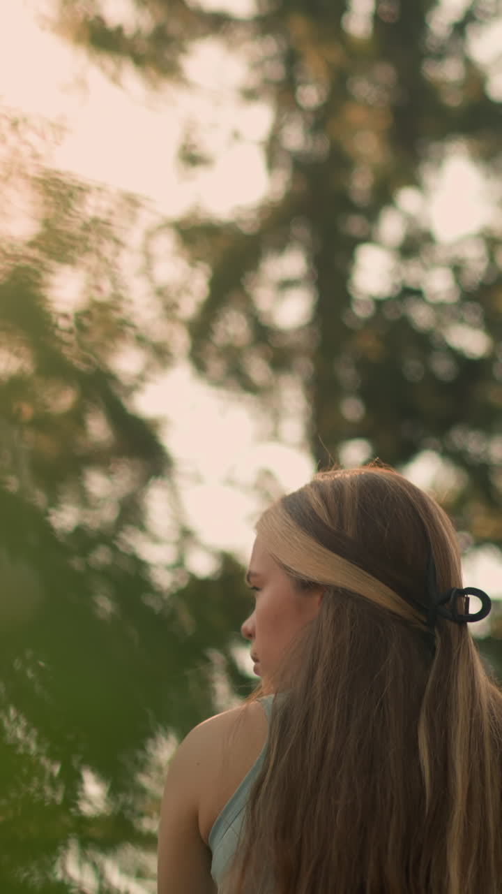 vista trasera de una joven con el cabello atado hacia atrás mirando pensativamente a la derecha mientras la luz del sol se filtra a través de las hojas, el fondo borroso con el suave efecto bokeh de los árboles y las luces lejanas