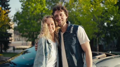 Portrait-of-a-beautiful-young-couple-blond-girl-and-brunette-guy-posing-against-the-background-of-a-gray-car.-Happy-couple-with