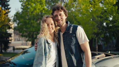 portrait of a beautiful young couple blond girl and brunette guy posing against the background of a gray car. happy couple with