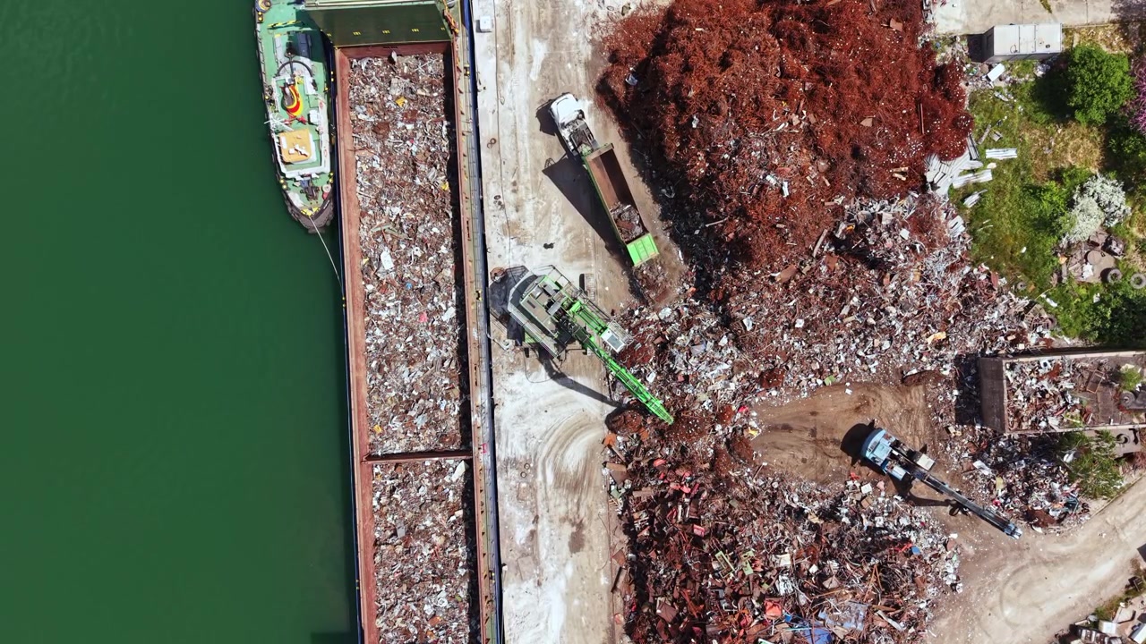 Attrezzature edili e gru lavorano in un deposito di rottami metallici vicino a uno specchio d'acqua durante le ore diurne in una trafficata zona industriale.