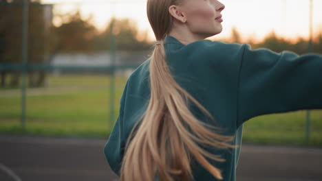 woman in green hoodie serving volleyball, flapping hand as ball rises, with open field and building in background, action shot of athlete training outdoors