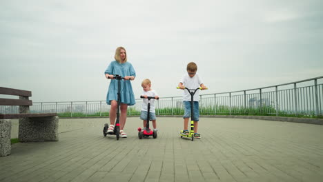 a mother and her two children ride scooters side by side on a paved outdoor path, the mother and older child each have their own scooter, while the younger child rides a smaller one