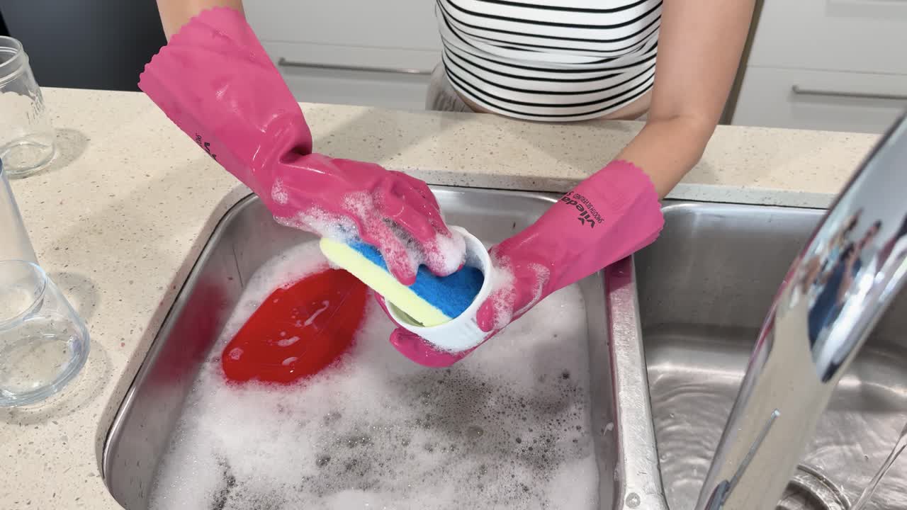 Person wearing pink gloves scrubs and rinses dirty dishes in a stainless steel kitchen sink under bright, even lighting with overhead camera angle