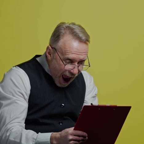 funny mature man with a folder isolated on yellow wall. happy old man in glasses writes on paper and depicts funny face. positive emotions.