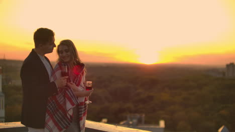 Young-married-couple-on-the-roof-hugging-and-drinking-red-wine-from-glasses-standing-dressed-in-plaid-and-admiring-the-beautiful-sunset-over-the-city..