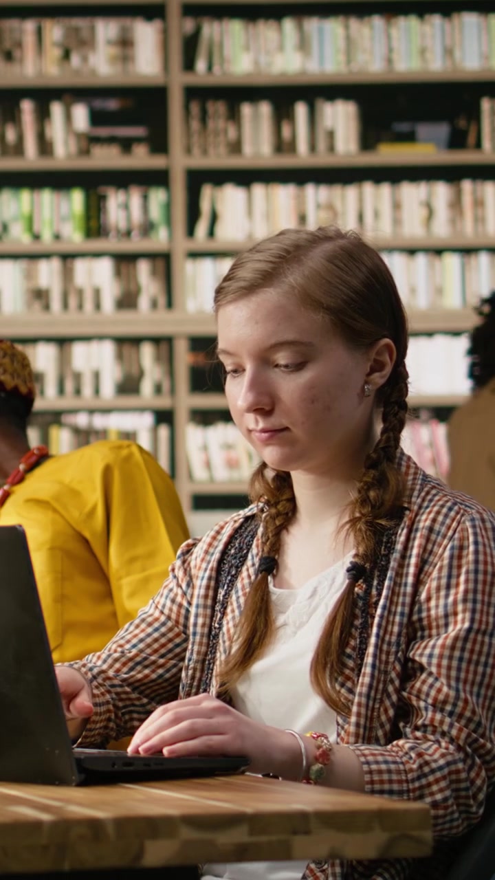 Vertikal video: Ung student arbeider på en bærbar datamaskin og tar notater ved et bibliotekbord med en åpen bok