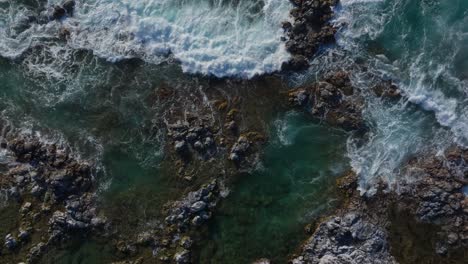 Strong-waves-crash-spreading-whitewash-across-rocky-shore,-drone-top-down-static