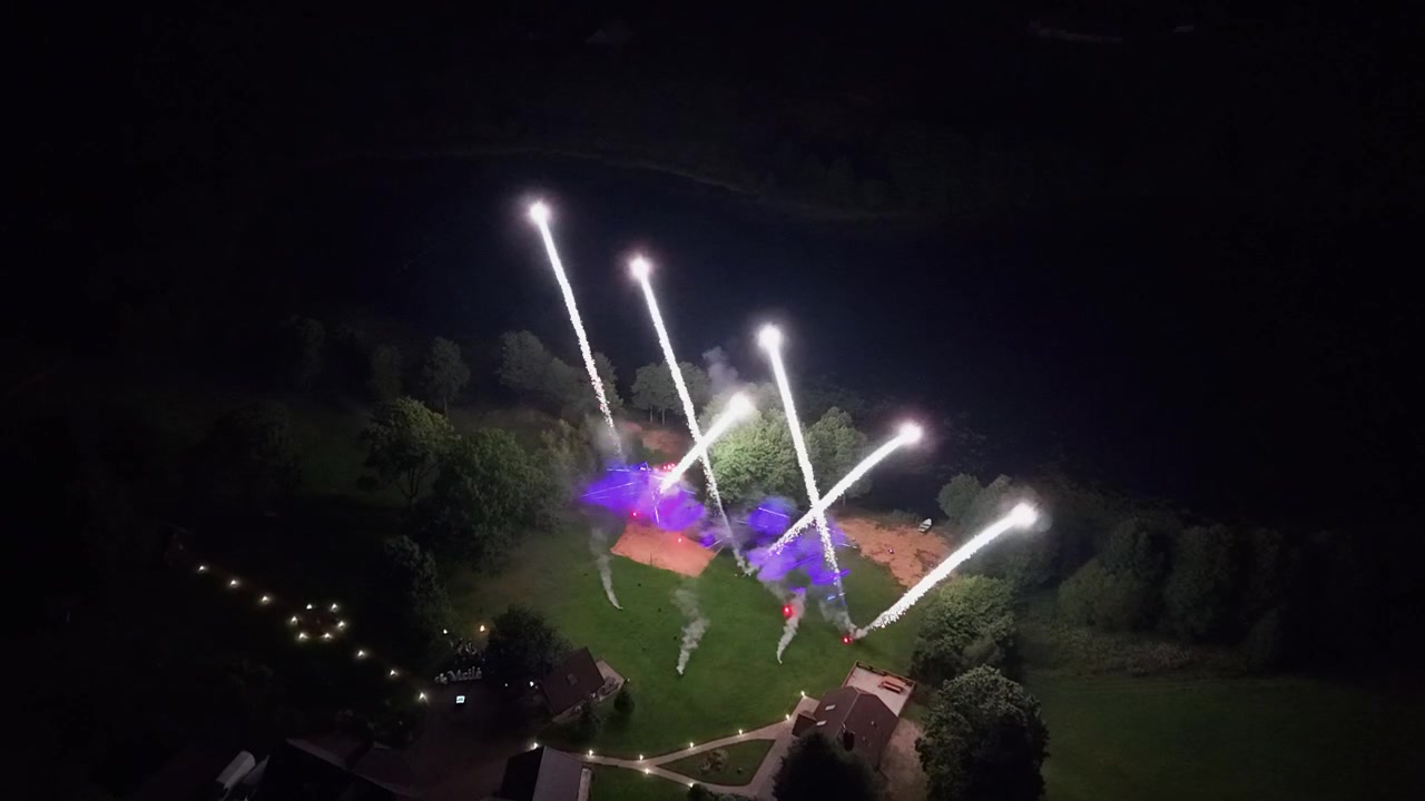 Dazzling drone video captures fireworks erupting above a lit park at night, with vibrant bursts of color and glowing pathways framed by surrounding greenery in a dynamic wide aerial shot.