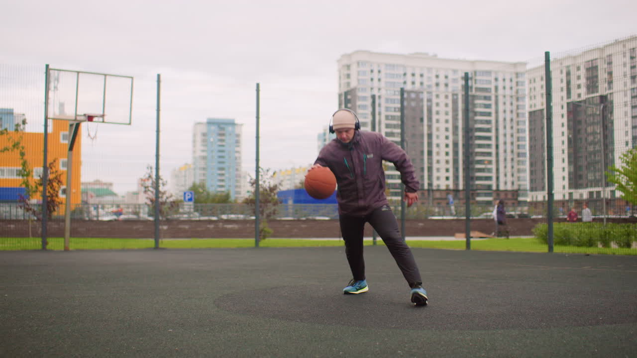 uomo che palleggia su un campo da basket all'aperto in un ambiente cittadino nuvoloso, con felpa e berretto, routine di riscaldamento intensa, alti condomini e recinzione metallica sullo sfondo, gioco di gambe costante e controllato