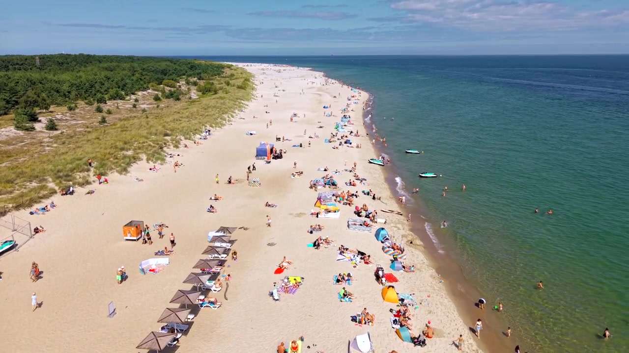 Imagem aérea de drone sobrevoando uma praia de areia lotada em Hel, na Península de Hel, na Polônia, com pessoas tomando sol e nadando no Mar Báltico turquesa, ao lado de uma costa arborizada em um dia ensolarado de verão.