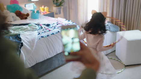 Niña-Feliz-Jugando-Con-Hula-hoop-Bailando-En-El-Dormitorio,-Madre-Tomando-Fotos-Usando-Un-Teléfono-Inteligente-Para-Compartir-En-Las-Redes-Sociales