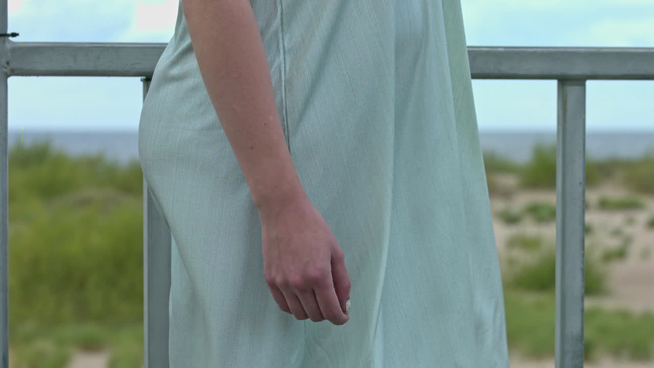 A pregnant woman standing in a vertical way at the balcony alone feeling the wind. An expectant female of childbirth having a quiet moment at the balcony. A pregnant woman wearing a loose dress