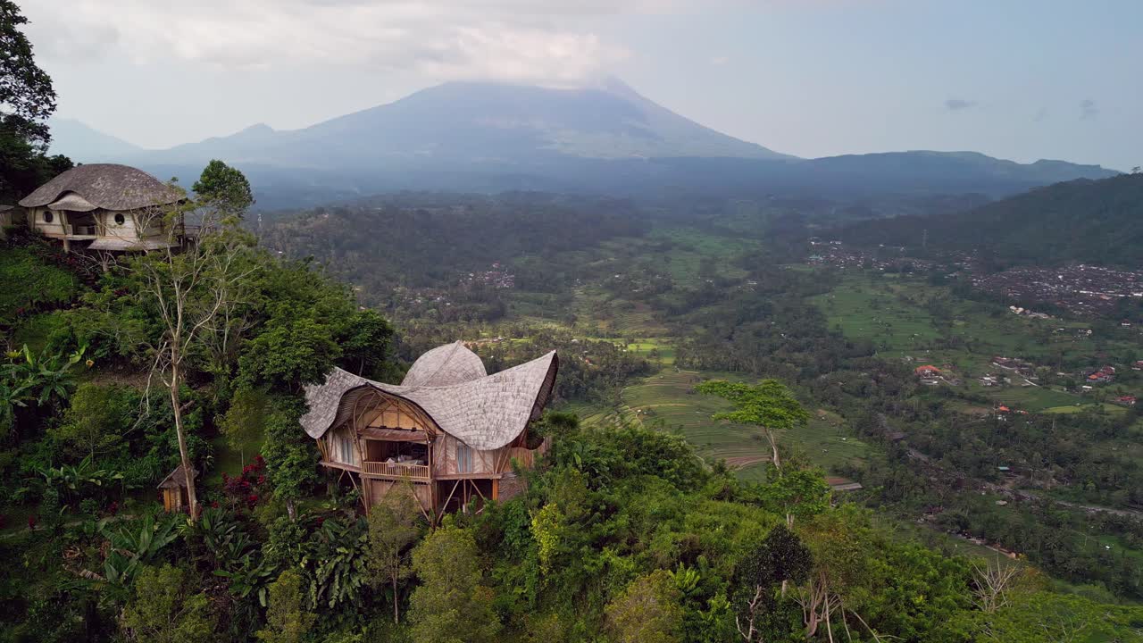 Peaceful aerial view of a bamboo house in the Sidemen highlands of Bali, surrounded by greenery, rice terraces, and the misty silhouette of Mount Agung in the distance