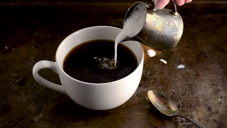 cup of coffee with thick cream being poured in slowly from a small metal creamer in dark setting