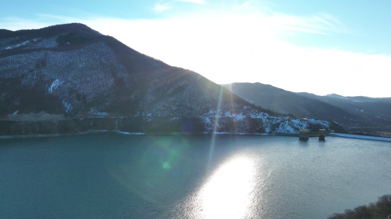 A winter lake reflecting the golden rays of the sun, bordered by wooded hills and mountains. The clear skies and calm water create a peaceful and inviting atmosphere