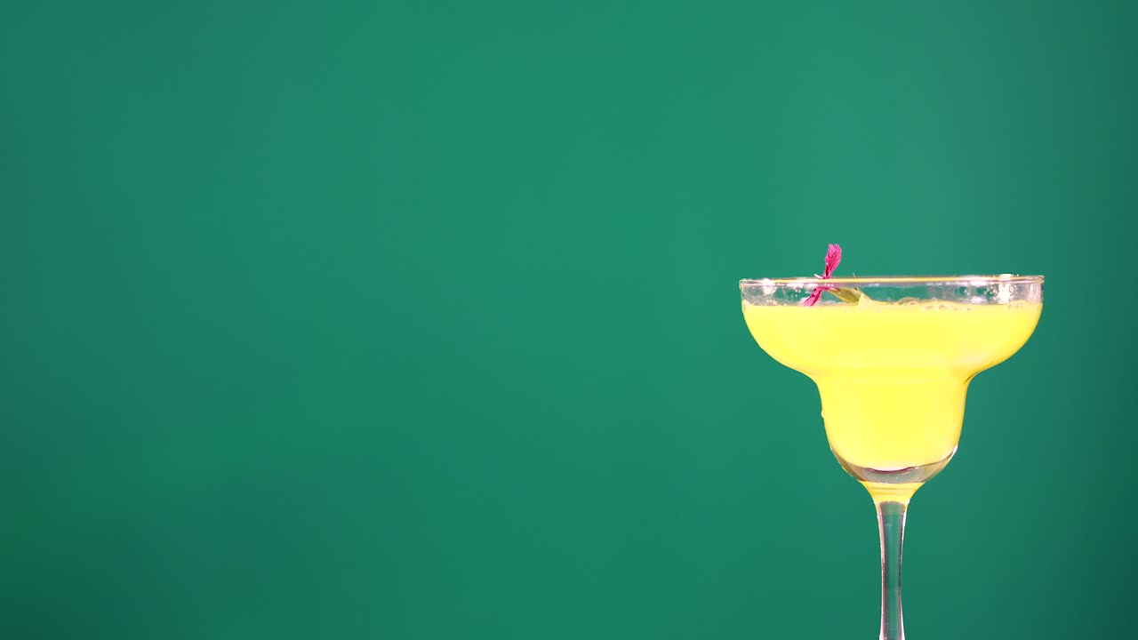 A yellow cocktail in a glass is garnished with a red cherry against a green background
