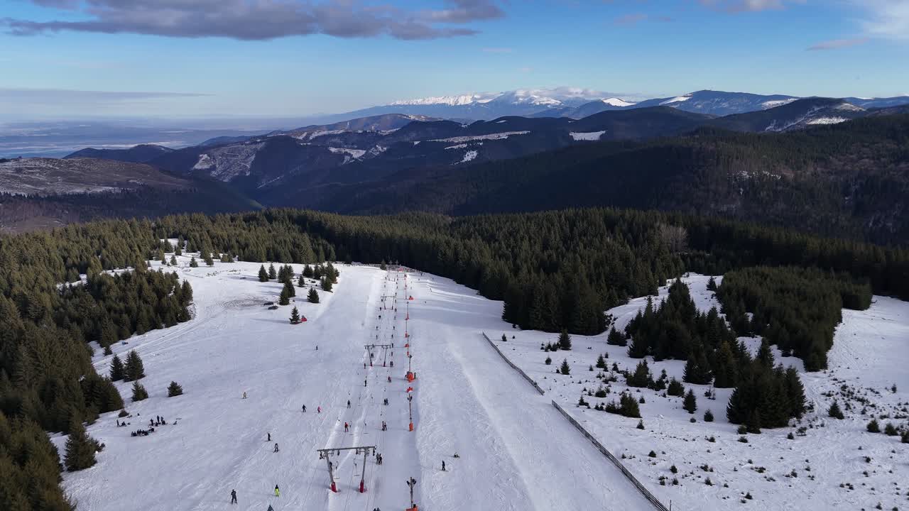 Letecké záběry z dronu lyžařského střediska v zimě, ukazující zasněžené svahy a lyžaře užívající si zimní sporty v horách. Malebná zimní krajina s horskými stromy, jasnou oblohou a aktivní turistikou.