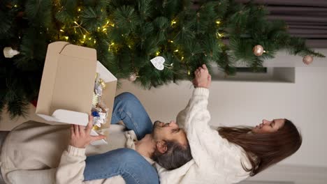 Vertical-video:-of-a-guy-holding-a-box-with-Christmas-decorations-in-his-hands-and-a-girl-sitting-on-his-shoulders-taking-decorations-from-the-box-and-attaching-them-to-a-Green-Christmas-tree