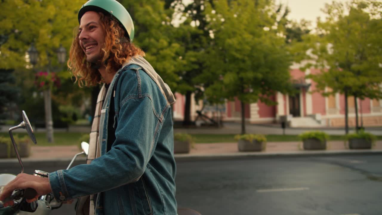 A happy brunette guy with curly hair in a denim jacket drives up on his moped, his girlfriend in a checkered shirt and a white helmet runs up to him, gets on the moped with the guy and they leave
