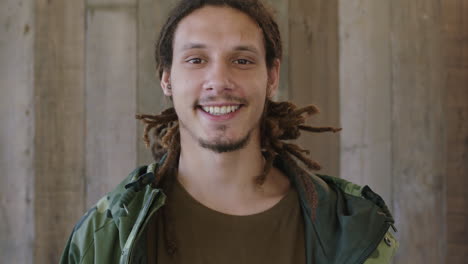 close up portrait of young attractive mixed race man dreadlocks hairstyle smiling looking at camera confident male wearing camouflage jacket wooden background