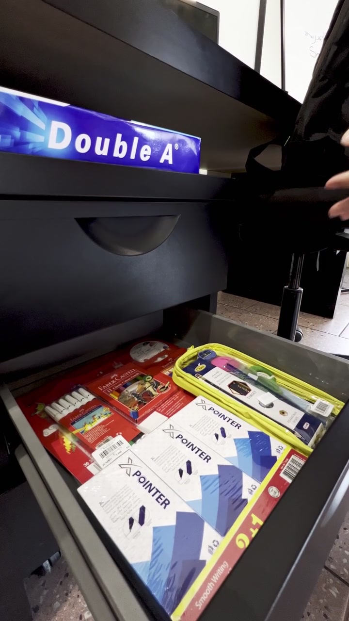 A close-up shot of a person's hand holding a box of pens over an open black office desk drawer. The modern indoor workspace setting features various colorful stationery supplies and copy paper.