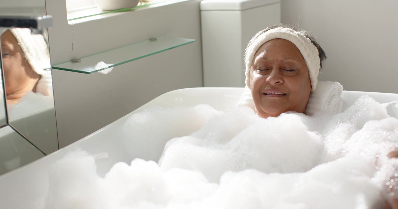 Donna afroamericana che si riposa nella vasca da bagno tra la schiuma, con un asciugamano arrotolato come fascia per capelli, mentre la telecamera si allontana