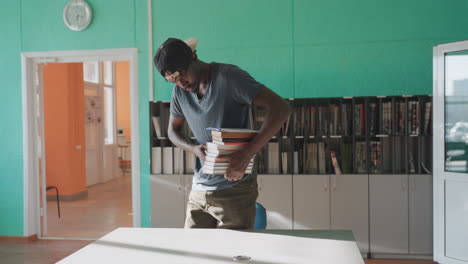 Student carrying stack of books places another on top of pile on table beneath teal wall and cabinets, wearing backward cap and grey shirt, focusing on neat arrangement in bright sunlit library room