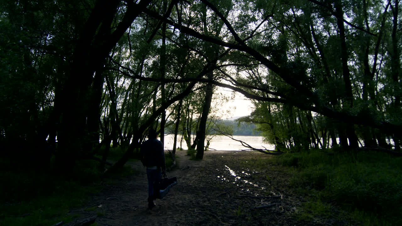 Man walking through a forest path towards a river at sunset