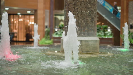 indoor water fountain with streams of water illuminated by colorful lights in modern shopping mall, surrounded by tiled pillar, escalator, and greenery