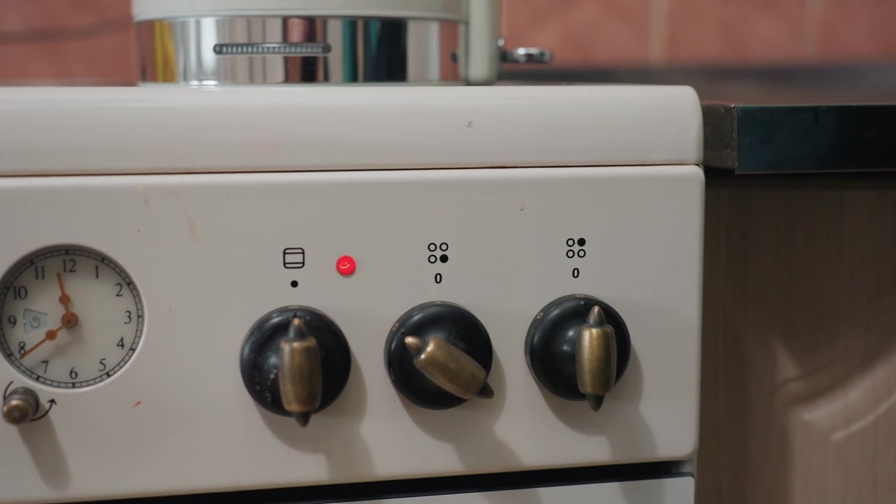 Nuovo inquilino che testa con cautela le manopole del fornello, primo piano di un fornello bianco vintage, indicatore rosso acceso, manopole nere in ottone, manometro meccanico, mano con maglione verde che regola la manopola, giorno del trasloco in un piccolo appartamento.