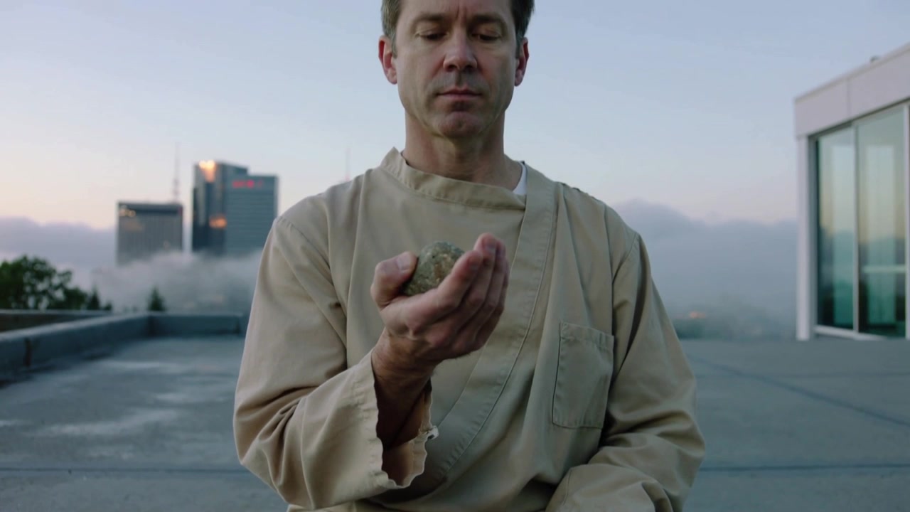 Afastando-se, a câmera revelando homem com roupão bege segurando pedra, meditando no telhado buscando calma