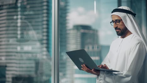 successful muslim businessman in traditional white kandura standing in his modern office, using laptop computer next to window with skyscrapers. successful saudi, emirati, arab businessman concept.