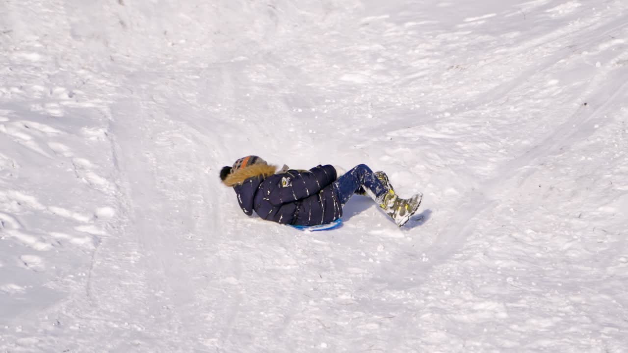 Cheerful kid on a snowy slide. Little boy on plastic sled riding down from white hill in winter. Cute child having fun at winter holidays. Slow motion.