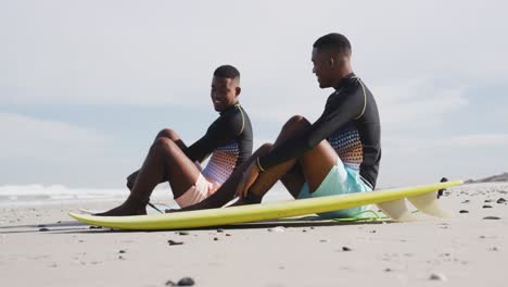 felices hermanos gemelos adolescentes afroamericanos sentados junto a las tablas de surf en una playa hablando