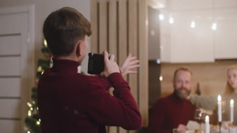 Un-Niño-Rubio-Con-Un-Suéter-De-Cuello-Alto-Rojo-Toma-Una-Foto-De-Sus-Padres-Y-Su-Hermana-Sentados-En-La-Mesa-En-La-Cena-De-Navidad