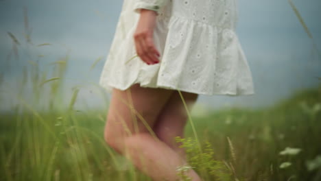 eine nahaufnahme des unterkörpers einer frau, während sie durch ein üppiges grünes feld geht und ein fließendes weißes kleid trägt. ihre hand hält sanft den mit perlen geschmückten rand des kleiders