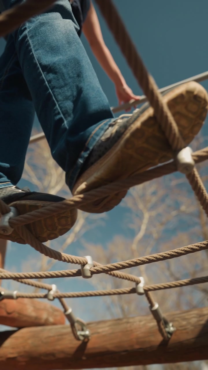 Primo piano di una persona che cammina con attenzione su un ponte di corda all'aperto con assi di legno e corde, indossando jeans blu e scarpe da ginnastica. La persona mantiene l'equilibrio sull'ostacolo elevato contro un cielo sereno.