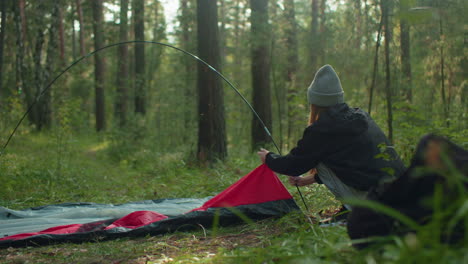 adventurer squats in dense forest holding flexible tent pole while placing tent fabric around base, sunlight glows gently through trees, adding warmth to tranquil outdoor setup moment