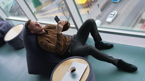 man rests in a soft chair with a phone. young man sitting near the window with a city transport background and using his mobile phone. cup of coffee on small table. top view.