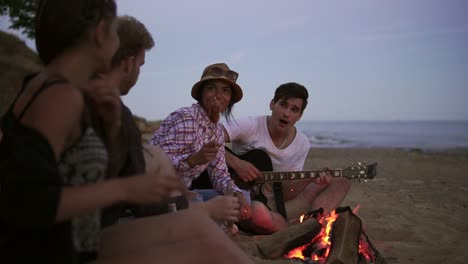 Picnic-of-young-people-with-bonfire-on-the-beach-in-the-evening.-Cheerful-friends-singing-songs-and-playing-guitar.-Slow-Motion